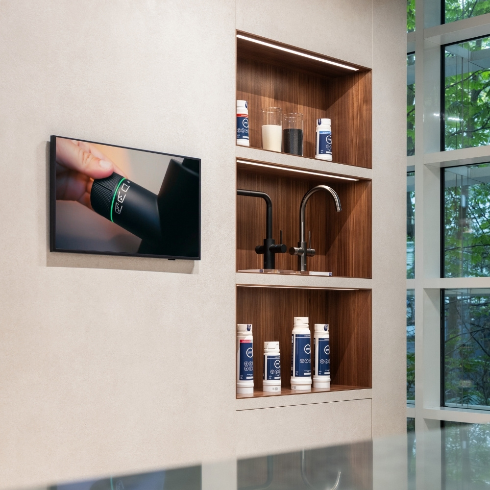 Wandmonitor neben beleuchteter Holznische mit GROHE Filterkartuschen und Armaturen, rechts Fensterfront mit Grün