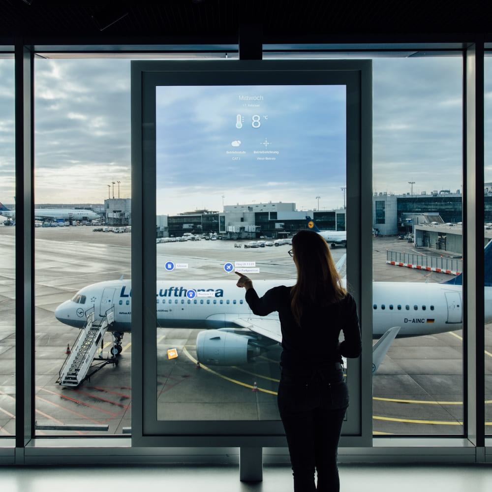 Passagier navigiert Flughafen-Touchscreen mit Wetter- und Fluginfo, Blick auf Lufthansa-Flugzeug