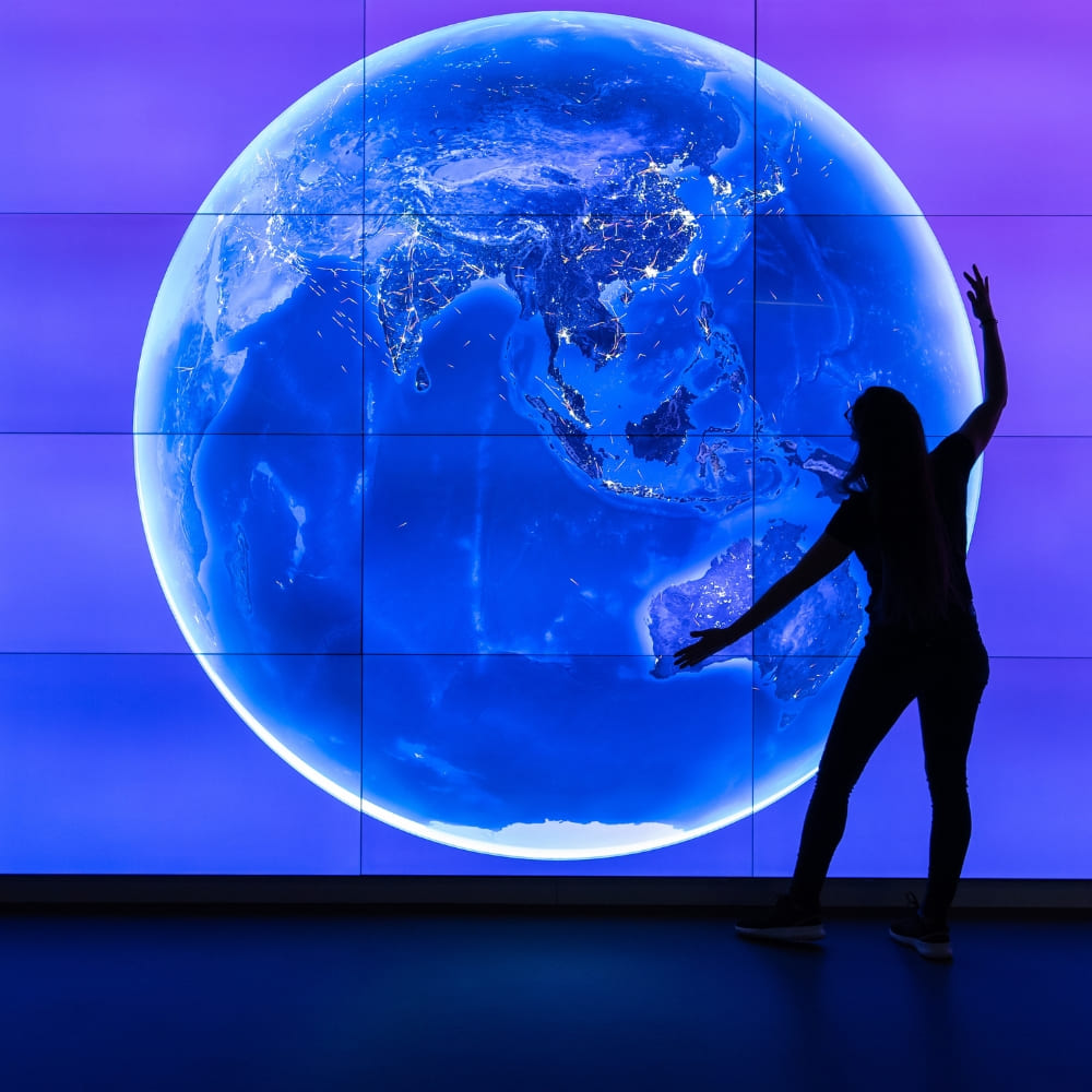 Große LED-Videowall zeigt leuchtende Weltkarte in Blau auf Lila Hintergrund, Person in Silhouette interagiert