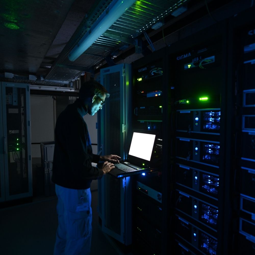  SIGMA-Techniker arbeitet an einem Laptop vor geöffneten AV-Equipment-Racks in einem abgedunkelten Technikraum mit blauer und grüner LED-Beleuchtung.